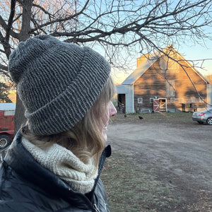 Person wearing a gray knit hat and white scarf standing in front of a rustic wooden barn.