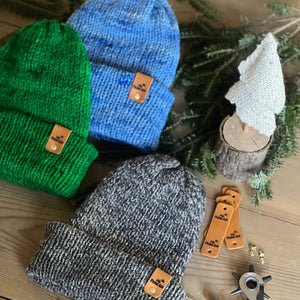 Three knit hats in green, blue, and gray with brown labels on a wooden surface.